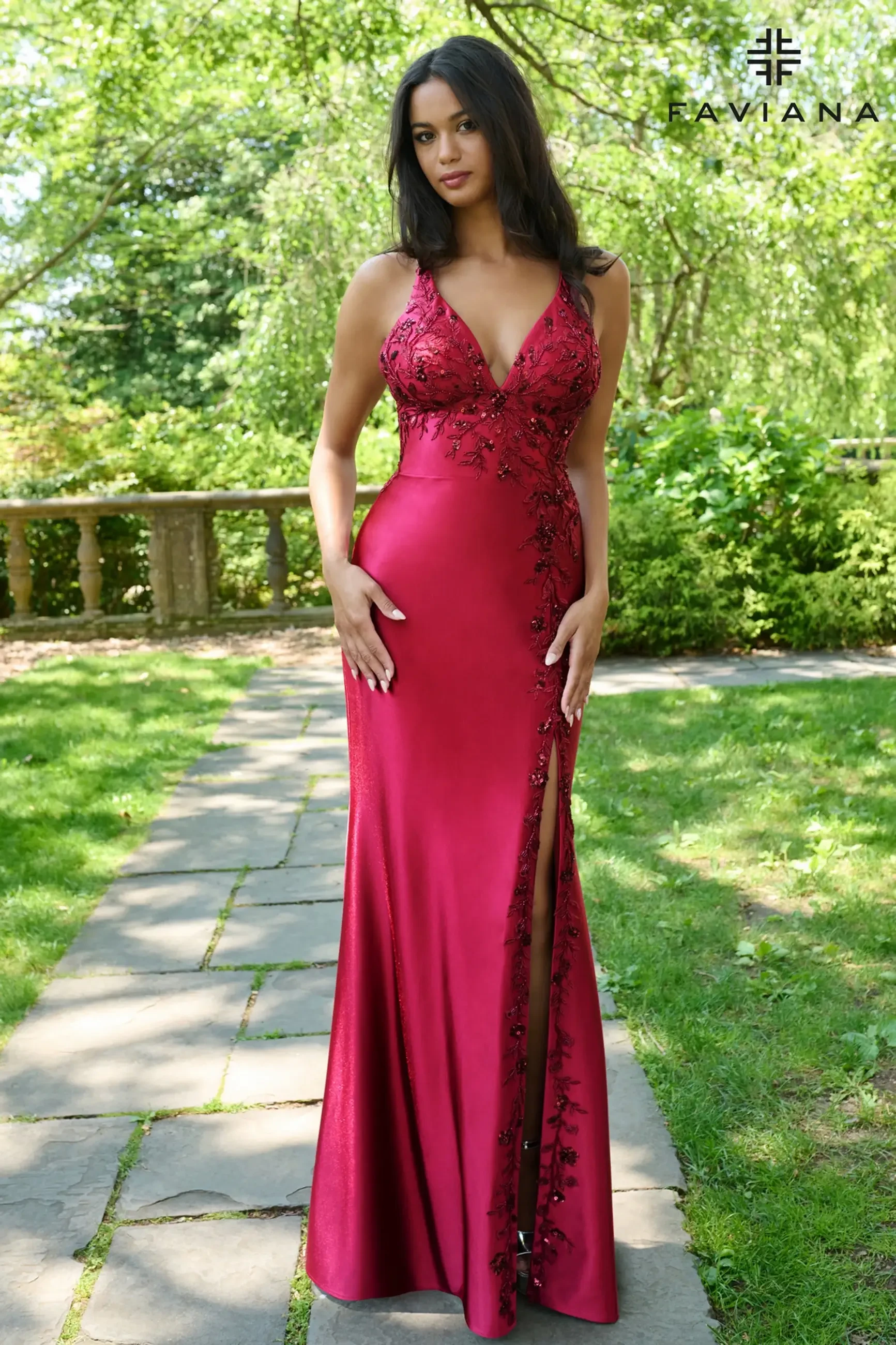 A woman stands outdoors wearing a fitted, deep red gown with floral embroidery and a thigh-high slit. The garden background is lush and sunlit.