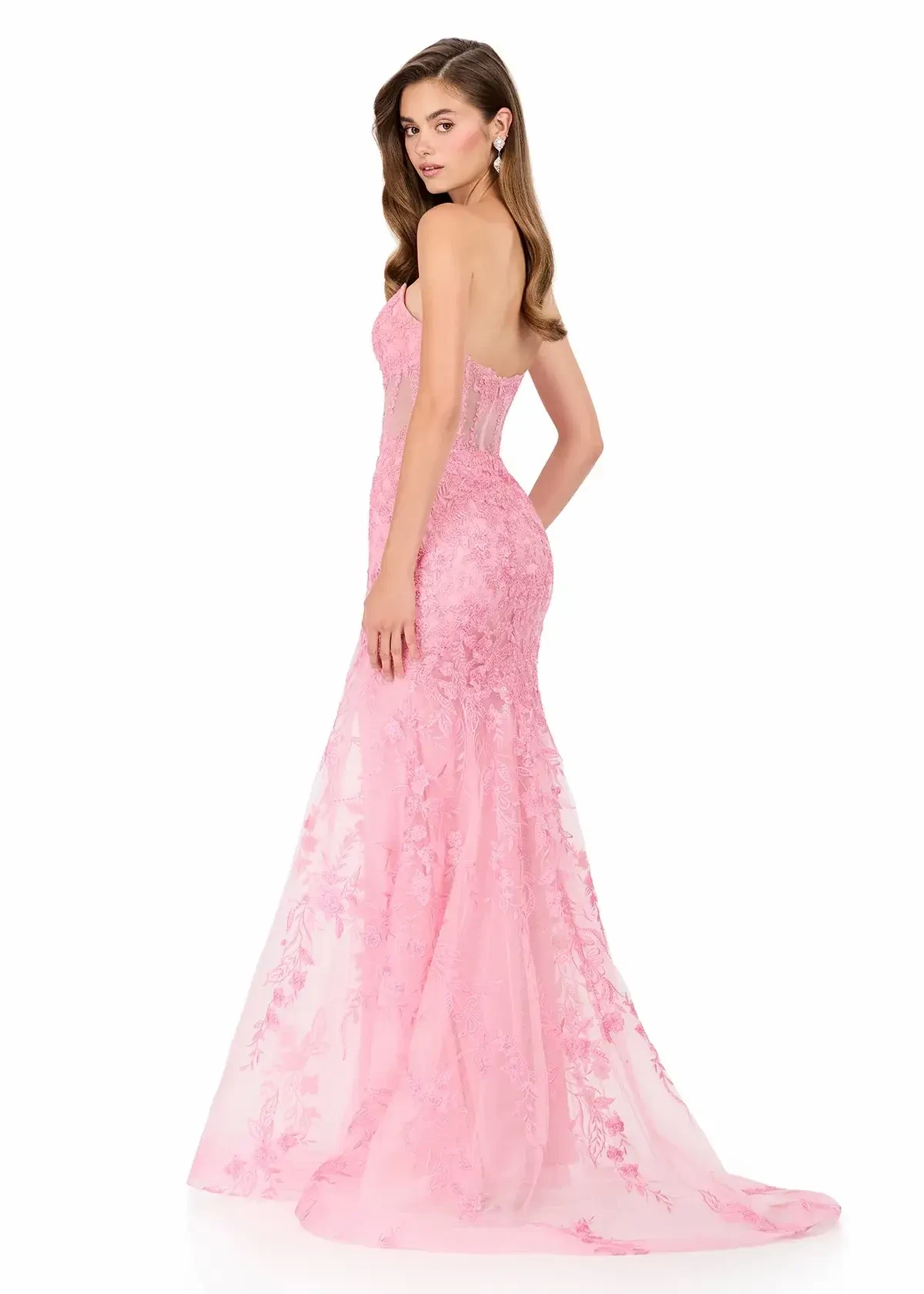 A woman in a strapless, pink lace dress poses elegantly from the side against a white background. The long gown features floral patterns and exudes a graceful, formal tone.