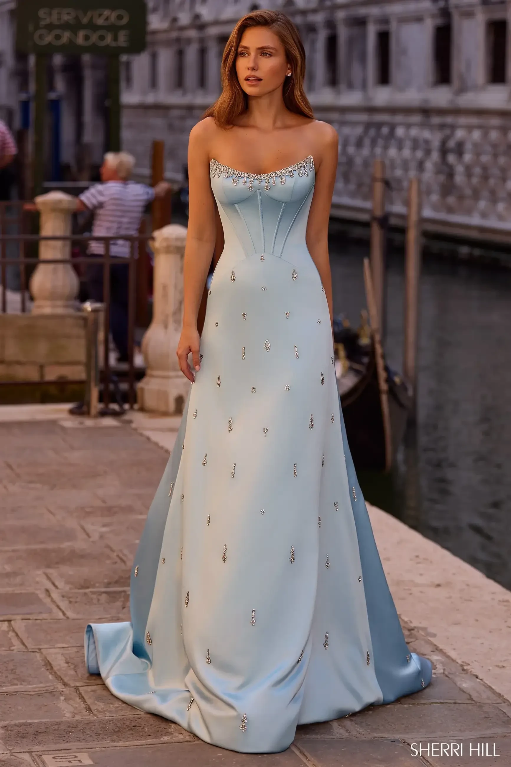 A woman wearing a strapless light blue gown adorned with sparkling embellishments, standing by a canal in a picturesque setting.