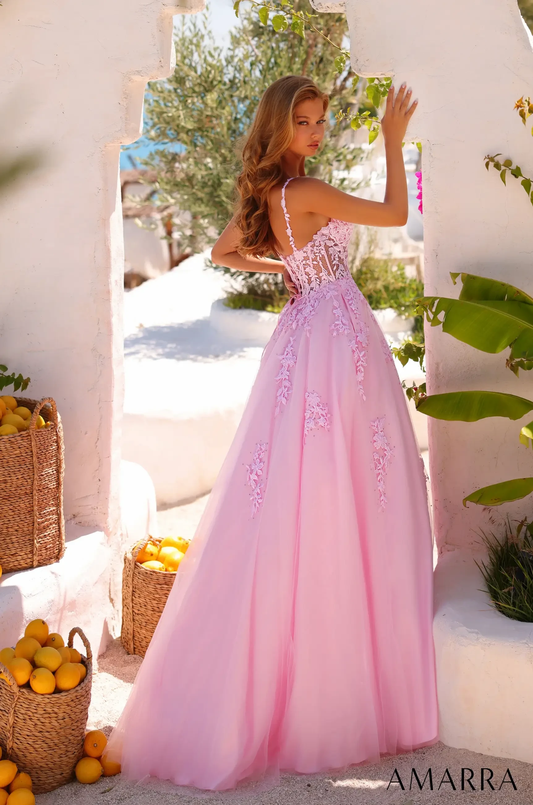 A woman in a flowing pink gown with lace details poses in a sunlit archway, surrounded by wicker baskets of lemons. The scene is serene and elegant.